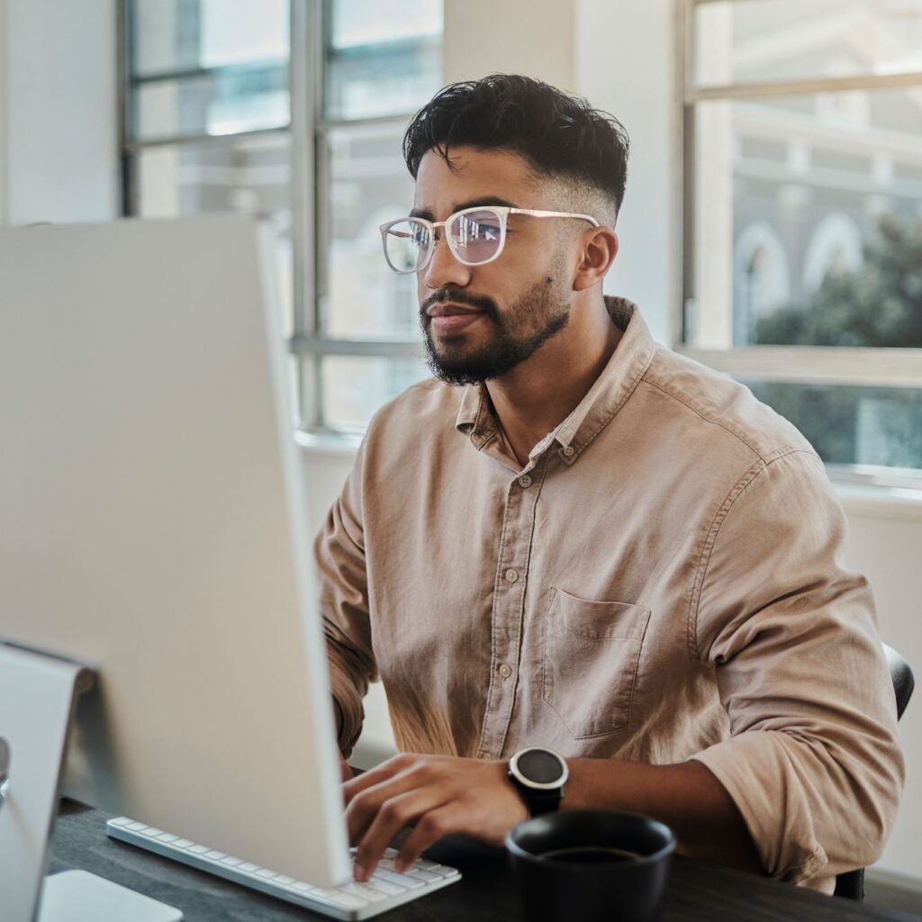 Man in glasses working on a computer at a desk with a coffee cup.