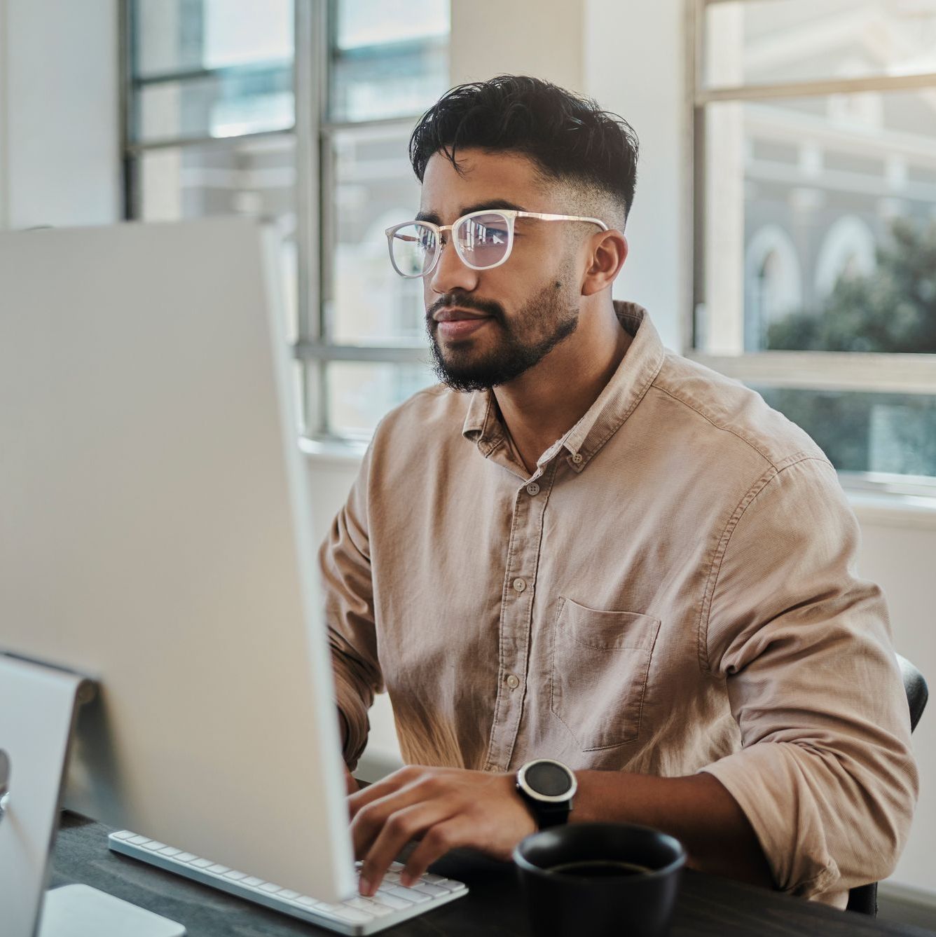 Man in glasses working on a computer at a desk with a coffee cup.